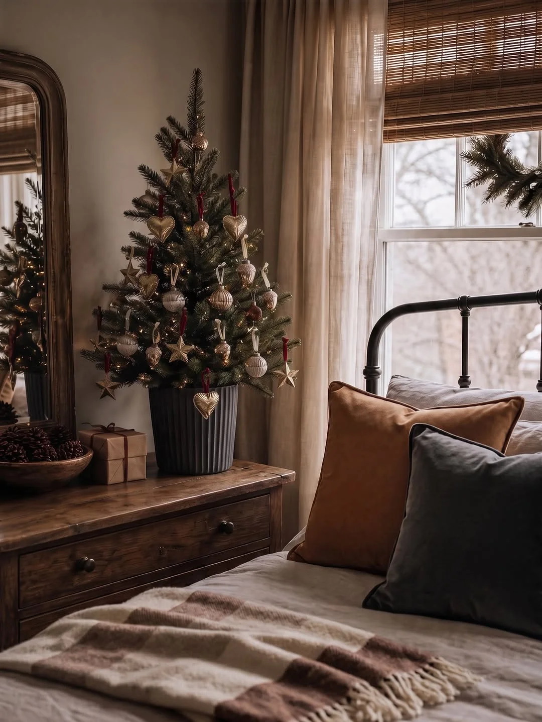 A bedroom corner with a nightstand holding a small decorated tree, pinecones and a gift, beside a bed with pillows and a throw