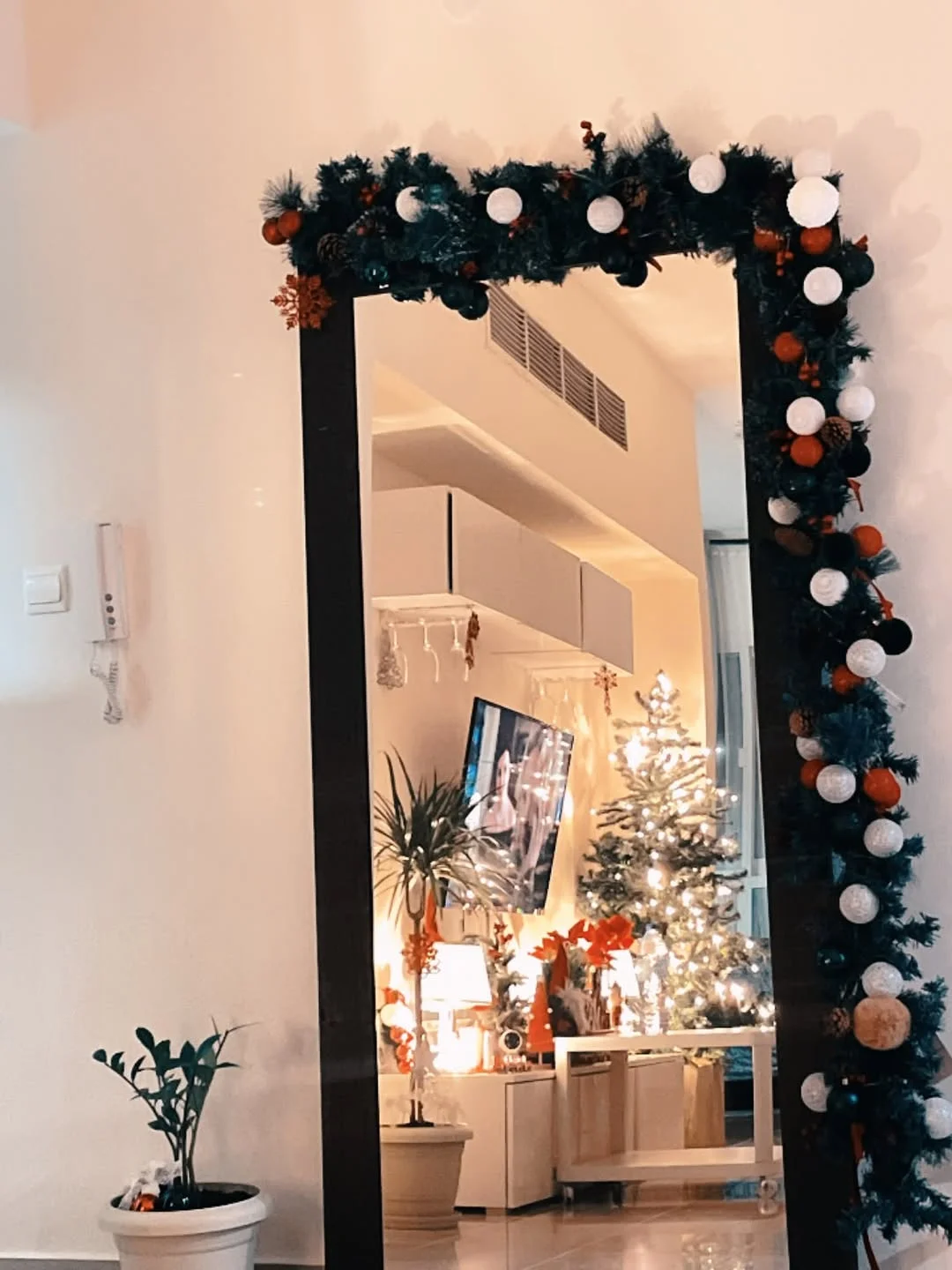 A tall floor mirror framed with dense green garland, reflecting a living area with a tree