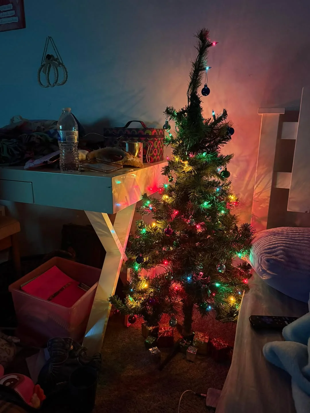 A desk corner with a small lit tree in a dim bedroom, with the edge of a bed visible